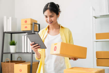 Startup small business, A young Asian woman checking online order on digital tablet and packing boxes for products to send to customers. working at the home office.の写真素材