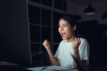 Happy excited Asian business woman celebrating success and working on computer at night.の写真素材