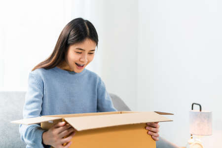Young Asian woman open box parcel while sitting on sofa.