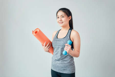 Smiley Asian woman wearing sportswear holding orange mat with blue dumbbell in living room. Healthy lifestyle concept.の写真素材