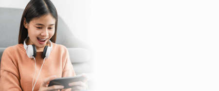 Excited young Asian woman wear white headphone on the head and playing games on smartphone in the living room at home.の写真素材