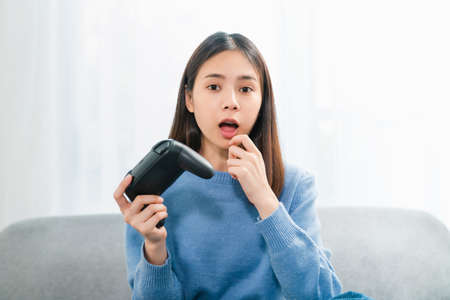 Excited young Asian woman sitting on sofa and holding joystick with playing games in the living room at home.の写真素材