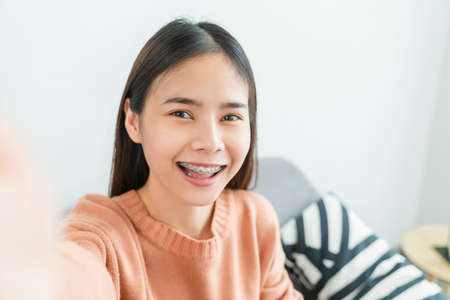 Cheerful beautiful Asian woman in a orange shirt and hand touching camera on sofa at house.の写真素材