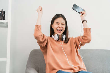 Excited young Asian woman wear white headphone on the head and playing games on smartphone in the living room at home.の写真素材