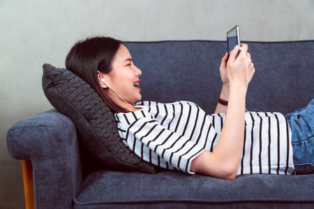 Happy young Asian woman holding a digital tablet and using online social lifestyle on sofa in the home. Technology for communication concept.の写真素材