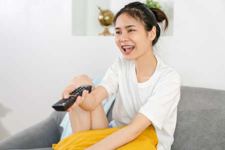 Young Asian woman sitting at the sofa in the house and holding the TV remote to watch movies on a relaxing day.の写真素材