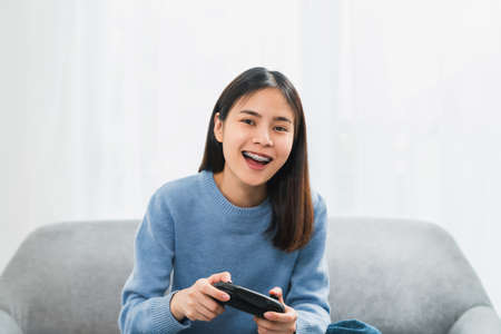 Excited young Asian woman sitting on sofa and holding joystick with playing games in the living room...