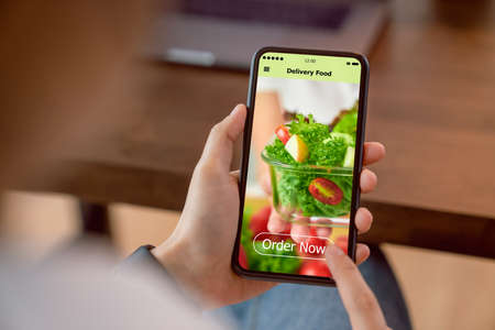 Woman using smartphone and touching application screen for ordering salad online on table at the home.の写真素材