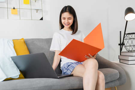 Happy young Asian woman working for laptop with graph documents from home on the sofa, meeting and...
