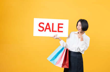 Young smiling asian woman holding multi coloured shopping bags and hands showing sale sign on light...