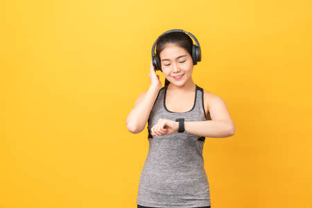 Smiley Asian woman wearing sportswear looking smartwatch and headphones on orange background.の写真素材