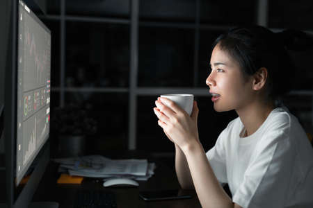 Asian businesswoman trader looking and drink coffee with graphs analysis candle line on table in...