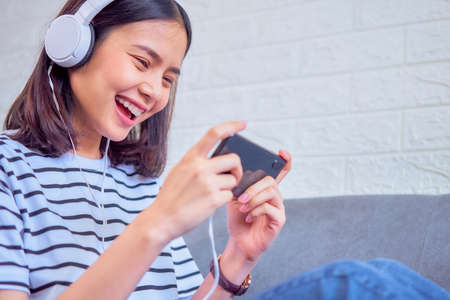 Excited young Asian woman sitting on sofa wear white headphone on the head and playing games on smartphone in the living room at home.の写真素材