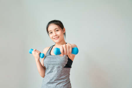 Smiley Asian woman wearing sportswear pumping up muscles with blue dumbbell in living room. Healthy lifestyle concept.の写真素材