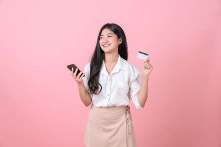 Young smiling Asian woman holding credit card with smartphone and looking to copy space on pink background.の写真素材
