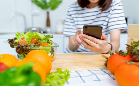 Woman hand holding a smartphone and salad bowl with tomato and various green leafy vegetables on the...