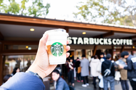 Tokyo, Japan - March 17, 2019: Hand holding Starbucks coffee cup with brand logo in front of Starbucks Coffee Ueno Onshi Park during pink cherry blossom or Sakura spring season of Tokyo, Japan.のeditorial素材