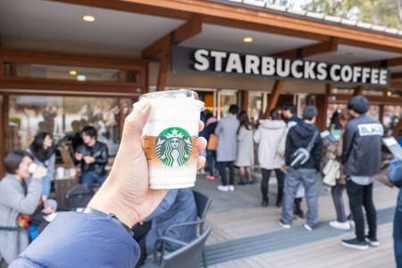 Tokyo, Japan - March 17, 2019: Hand holding Starbucks coffee cup with brand logo in front of Starbucks Coffee Ueno Onshi Park during pink cherry blossom or Sakura spring season of Tokyo, Japan.のeditorial素材