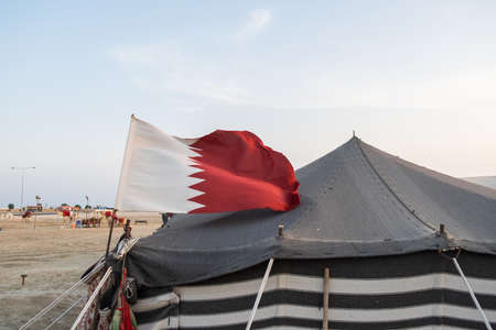 View of Qatar national flag with the camp at Desert Safari Camel Ride, a landmark for desert activities, in Al Wakrah, Qatar.の写真素材