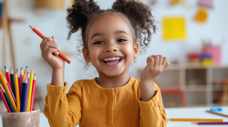 A young girl is smiling and holding a red pencil. She is in a classroom with a variety of colorful pencils and crayonsの素材
