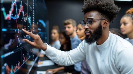 A man pointing at a computer screen with a group of people watching. The man is wearing glasses and has a beard. The people in the background are looking at the screen with interestの素材