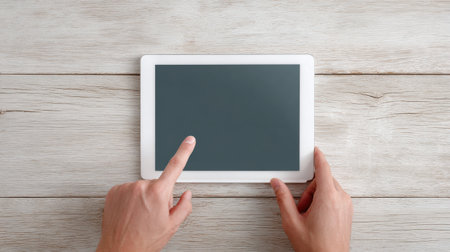 A person is holding a tablet with a black screen and pointing at it. The tablet is on a wooden tableの素材