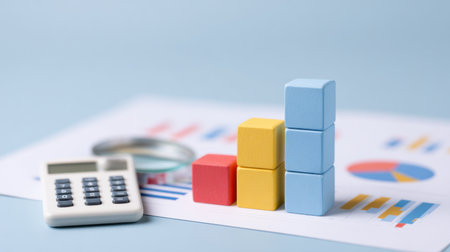A calculator, a stack of cubes, and a graph paper are on a table. The calculator is on the left side of the cubes, and the cubes are on top of the graph paper. The cubes are of different colorsの素材