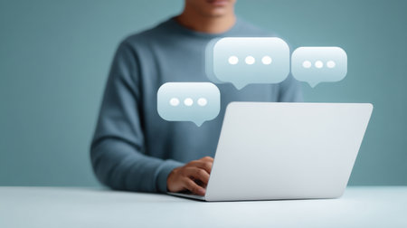A man is typing on a laptop with a blue shirt on. The laptop screen shows three speech bubbles with the words "chat" on themの素材