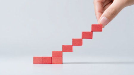 A hand is holding a stack of red blocks, which are arranged in a staircase pattern. The blocks are made of a material that resembles sugar, and they are placed on a white surfaceの素材