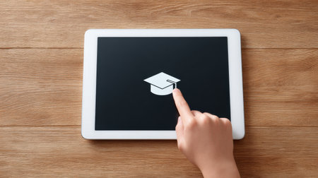 A tablet with a graduation cap on it. A finger is pointing at the graduation cap. The tablet is on a wooden tableの素材