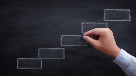 A hand is drawing a chalkboard with a series of squares on it. The squares are stacked on top of each other, creating a staircase-like structure. Concept of progress and growthの素材