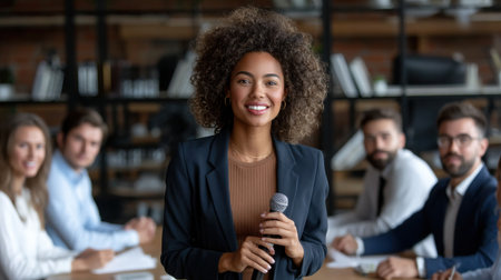 A woman is standing in front of a group of people, holding a microphone. She is smiling and she is confident. The group of people are seated around her, and there are several booksの素材