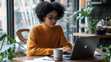 A woman wearing glasses is sitting at a table with a laptop and a cup of coffee. She is working or studying, as she is focused on her laptop. The scene suggests a quiet and focused atmosphereの素材