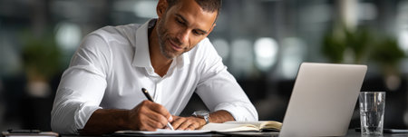 A man is sitting at a desk with a laptop and a notebook. He is writing on the notebook and using a pen. The scene suggests that he is working on a project or taskの素材