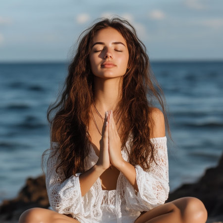 A woman is sitting on a rock by the ocean, praying and looking out at the water. She is wearing a white dress and has her hands clasped together in front of her chest. Concept of peace and tranquilityの素材