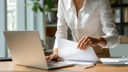 A woman is sitting at a desk with a laptop and a stack of papers. She is looking at the papers and seems to be focused on her work. Concept of productivity and concentrationの素材