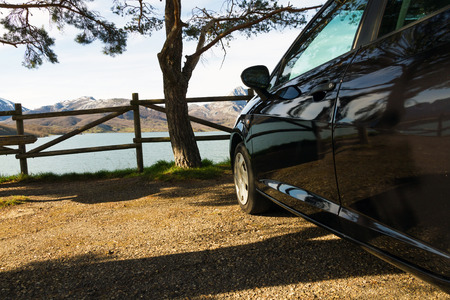 Side black car, parked in a rest area or lookout road, in the shade of a tree and a fence of wooden posts. Against a backdrop of mountains and lake or swampの写真素材