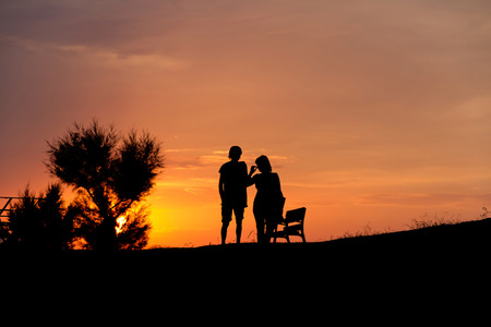 Silhouettes of a couple of girl and boy, backlit, in a park with the sun at sunset behind a bush and sky tinged With warm tones of fire. Next to a bank in the parallel drive to the coast in Llanes Asturiasの写真素材