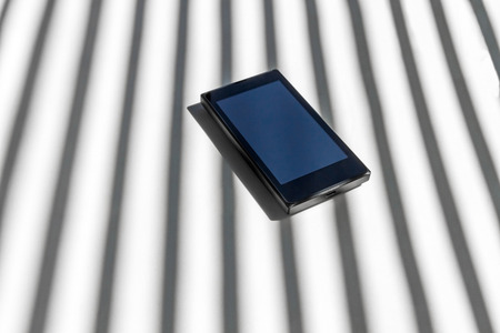 Mobile smartphone on white background. Between sunlight and shadow lines or bars. With input connections in the foreground - Telefono movil smartphone sobre fondo blanco. Entre la luz del sol y barrotes o lineas de sombra. Con la entrada para conexiones eの写真素材