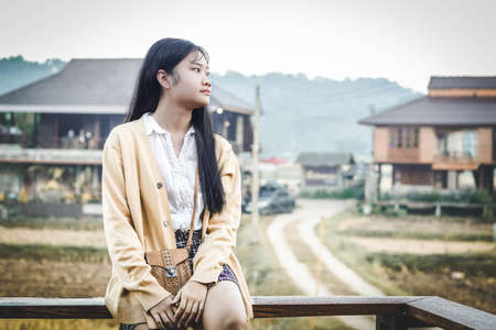 a girl is sitting on long bench in front of countryside houses in morningの写真素材