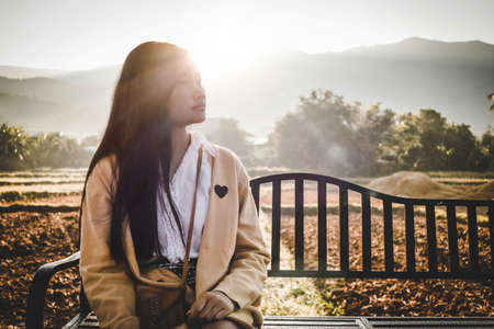 a girl is sitting on a bench among countryside field in an autumn morningの写真素材
