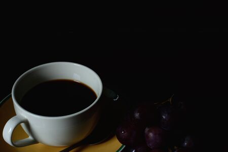 Black coffee in white cup with red grapes, isolated on black background.の写真素材