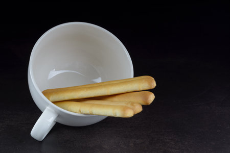 Bread sticks in white coffee cup on natural dark background.