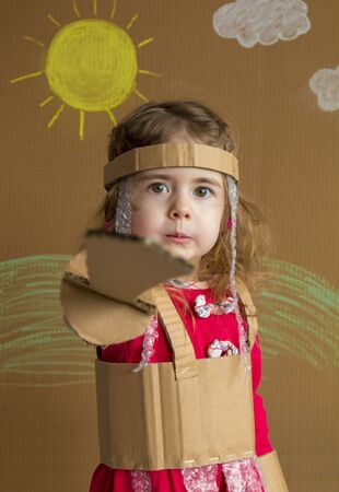 Clouse-up portrait of cheerful little girl with a cardboard sword and a suit. handmade. background of painted sun and white cloudsの写真素材