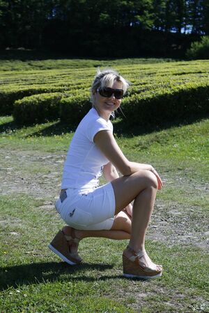 Cute blonde in a white T-shirt and shorts sits on the nature of the summer on a background of green treesの写真素材
