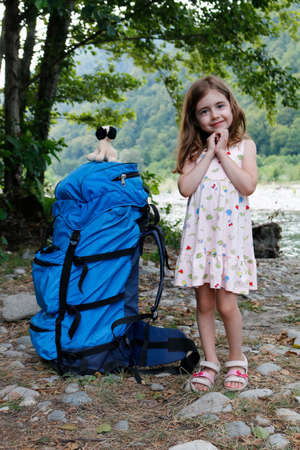 A large backpack for an adult stands next to a little girl against the backdrop of trees and a river, on a sunny day.の写真素材