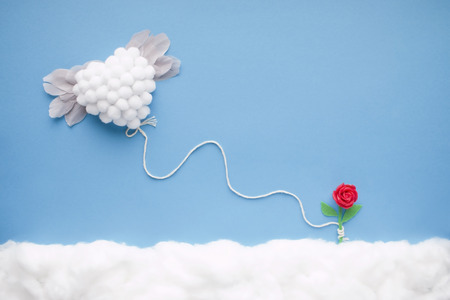 Flying white heart with feather wings, attached by a white string to a tiny handmade red flower, in a heavenly scenery with clouds made of cotton-wool, on a blue sky paper background.の写真素材