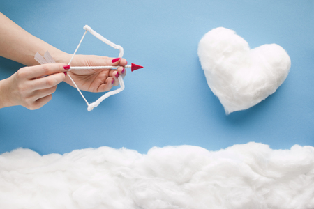 Two hands shooting an arrow to a cotton-wool heart, in a heavenly scenery with cotton wool clouds on a blue sky paper background.の写真素材