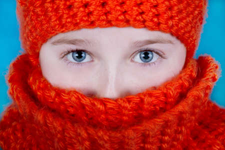 A close up of an attractive girl with blue eyes, wearing an orange winter hat and scarf. Against a blue background. Features the complimentary colors orange and blue.の写真素材