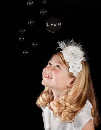 A cute girl wearing a white dress and white headband looking up at bubbles. With a black backgroundの写真素材